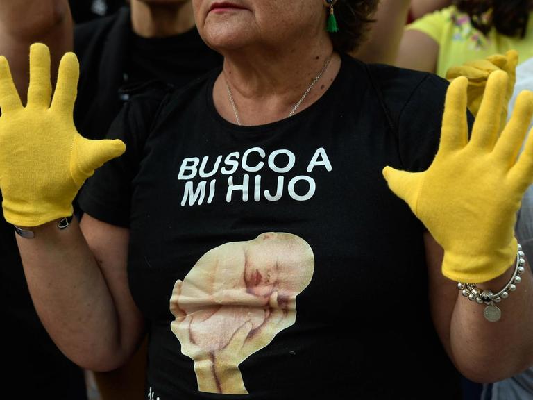 Eine Frau trägt ein schwarzes T-Shirt mit einem aufgedruckten Baby-Foto. Darauf steht: "Ich suche meinen Sohn. Geboren im April 1976 in Cádiz."