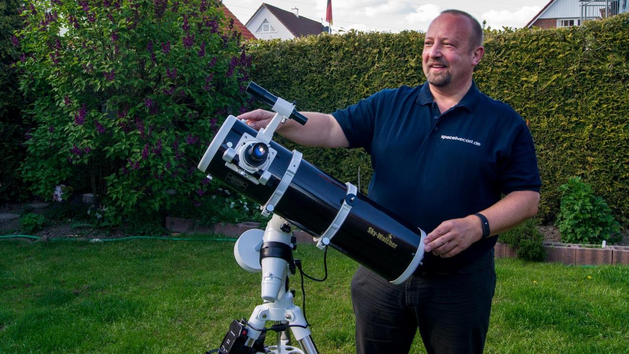 Amateurastronom Thomas Wehr steht hinter seinem Teleskop im Garten. Hinter ihm ist eine sauber geschnittene Hecke und dahinter Einfamilienhäuser.