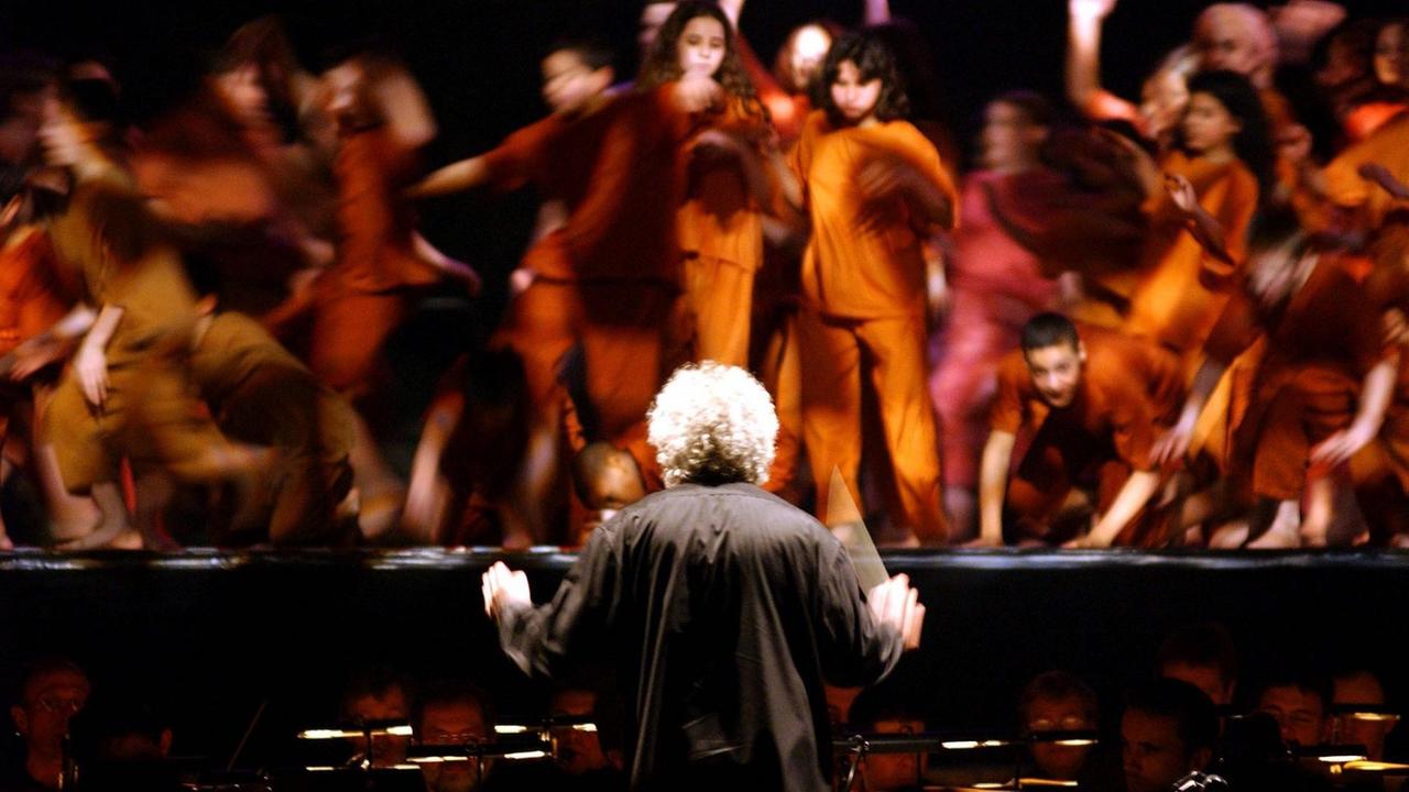 Der Dirigent ist von hinten mit seinen weißen Locken zu sehen, wie er vor wirbelnden Tänzern dirigiert, die in rot-braunen Kostümen agieren.
