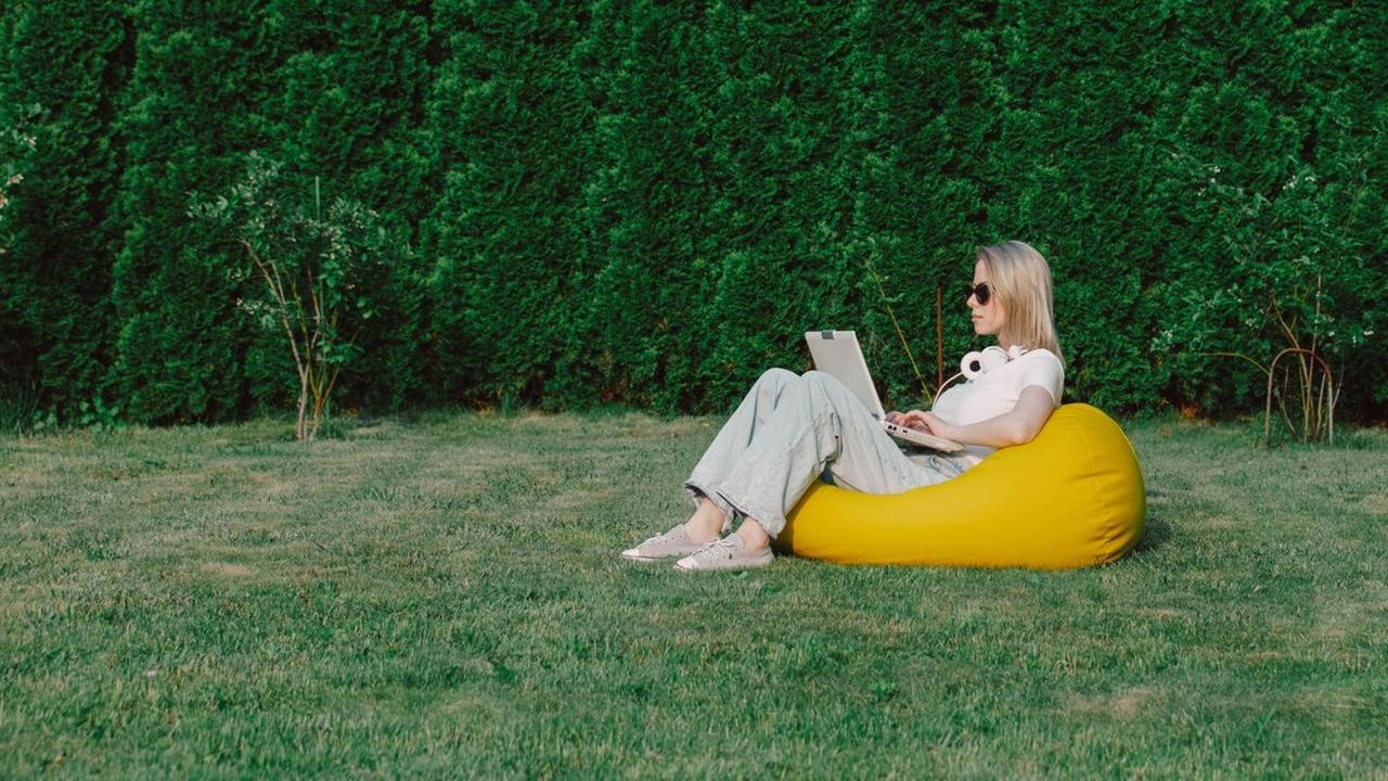 Eine Frau sitzt mit ihrem Laptop auf einem gelben Sitzsack auf einer Wiese vor Bäumen
