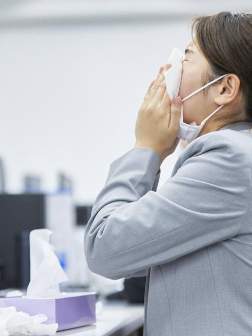 Eine Frau sitzt mit Maske an ihrem Büroarbeitsplatz und niest.