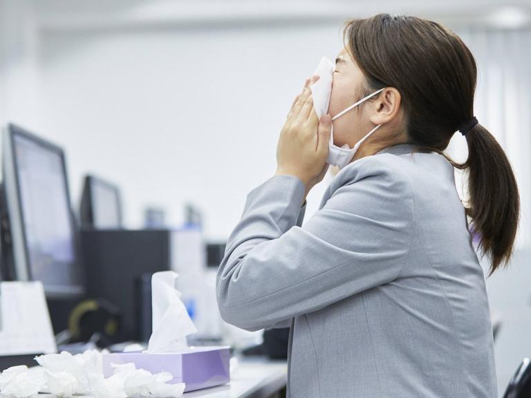Eine Frau sitzt mit Maske an ihrem Büroarbeitsplatz und niest.
