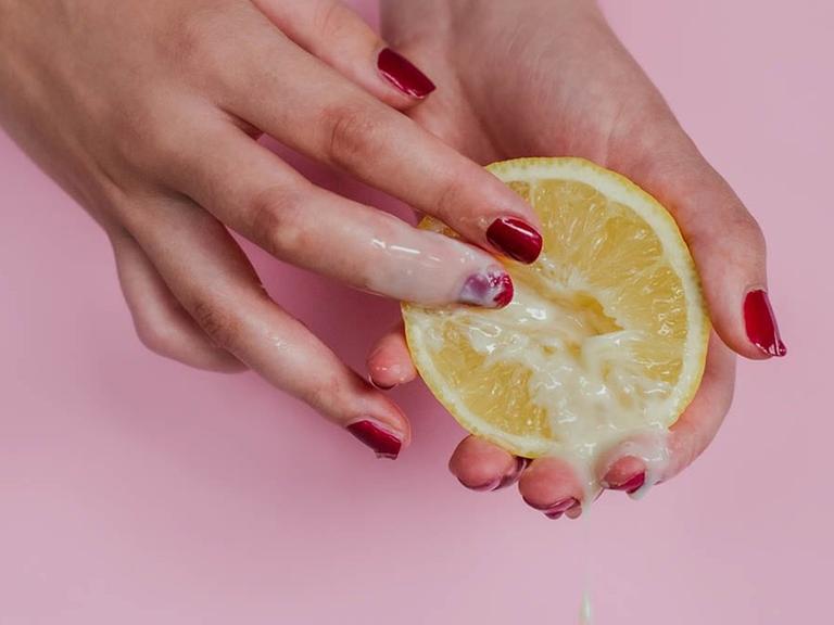 Vor rosa Hintergrund halten zwei Hände mit rotem Nagellack eine Zitronenhälfte, über die eine milchige Flüssigkeit läuft.