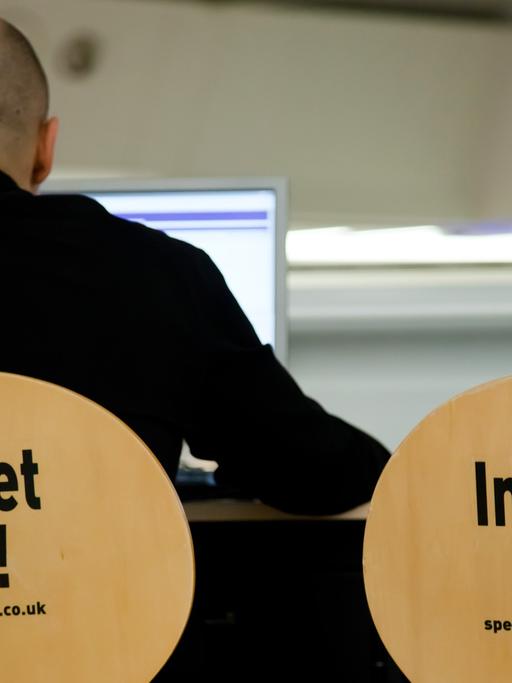 Ein Mann sitzt auf einem Stuhl, auf dessen Rückenlehne "Internet here!" steht, neben ihm ein identischer Stuhl und ein Computerbildschirm