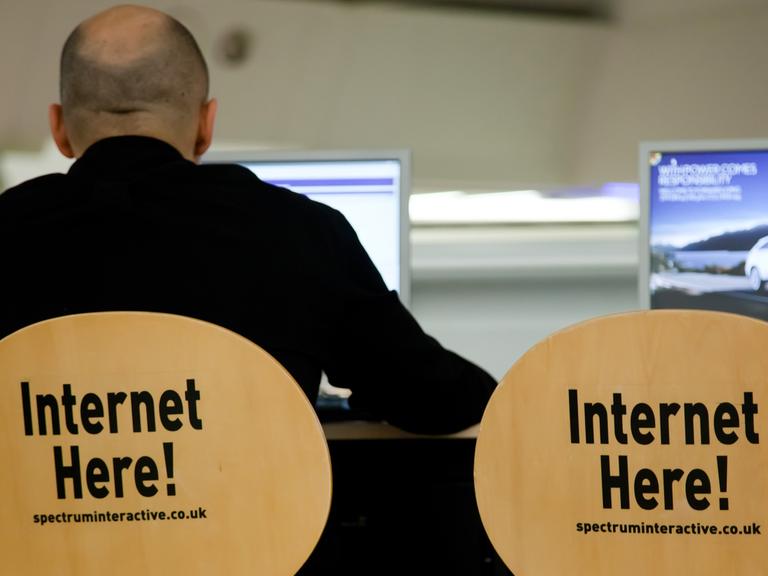 Ein Mann sitzt auf einem Stuhl, auf dessen Rückenlehne "Internet here!" steht, neben ihm ein identischer Stuhl und ein Computerbildschirm