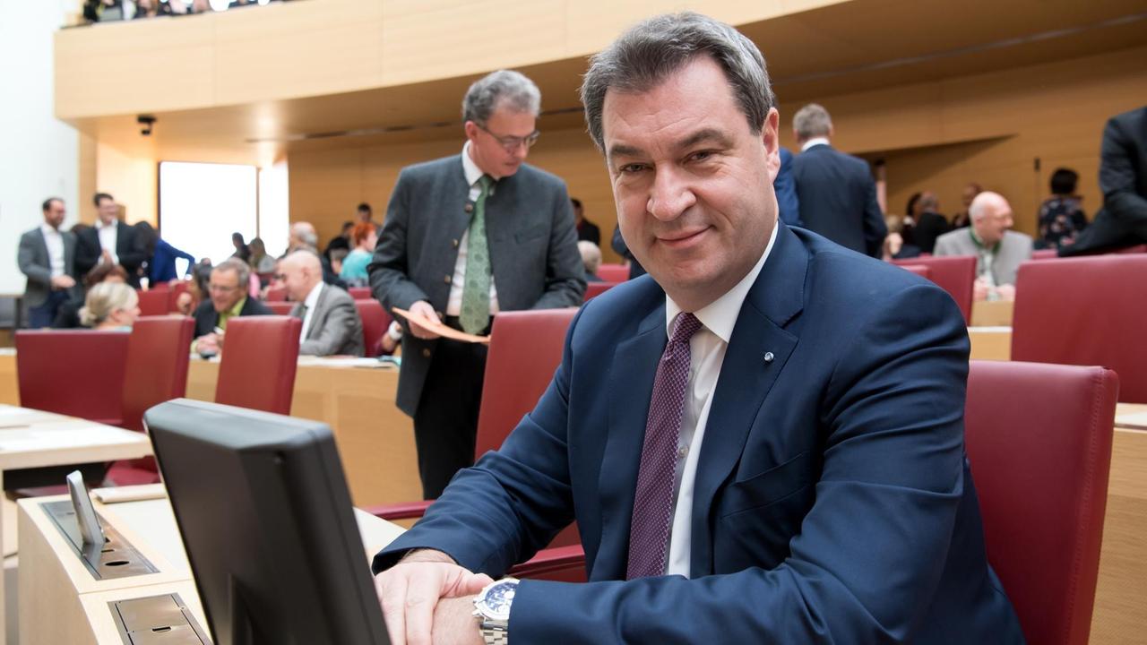Söder sitzt auf seinem Platz im Landtag und lächelt in die Kamera.