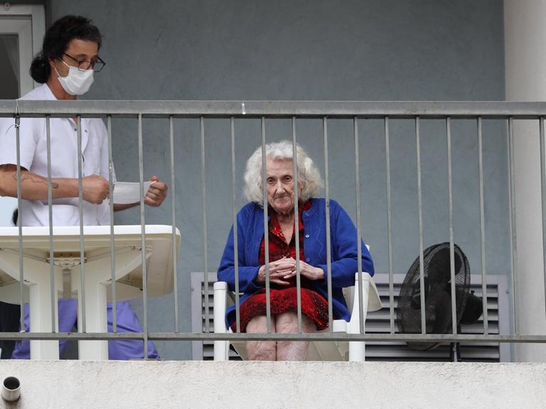 Auf einem Balkon eines Pflegeheims sitzt eine ältere Dame, Neben ihr steht eine Pflegerin mit Mundschutz.