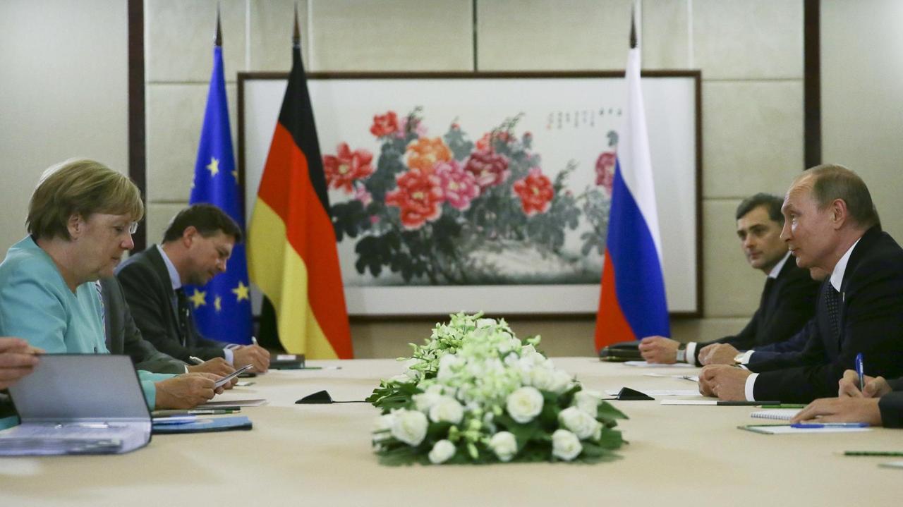 Merkel und Putin sitzen sich mit Mitarbeitern an einem Tisch gegenüber.