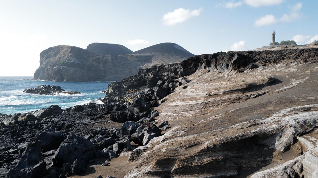 Eine schwarze hügelige Vulkanlandschaft ohne Vegetation am Meer.