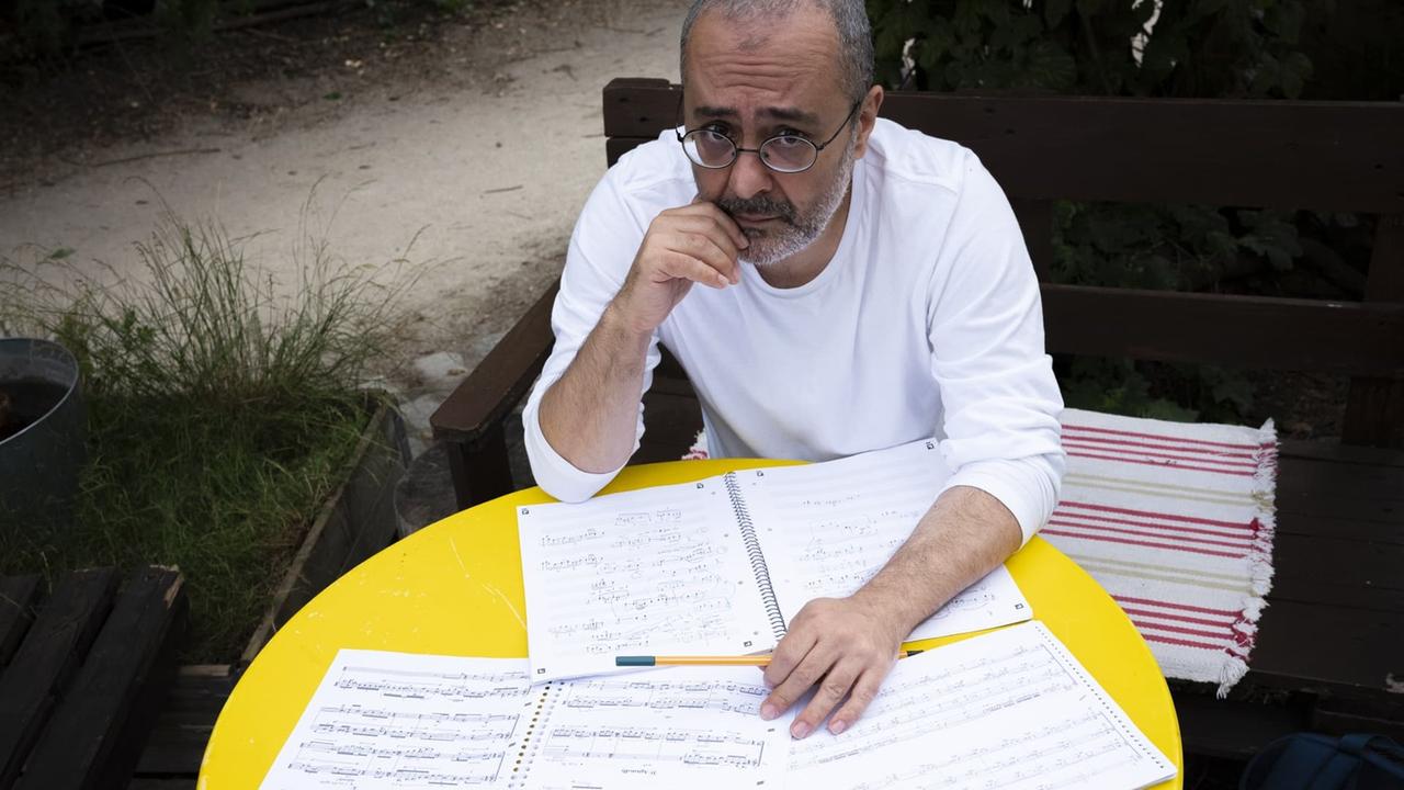 Ein Mann mit Brille sitzt an einem gelben, runden Gartentisch voller Notenblätter.