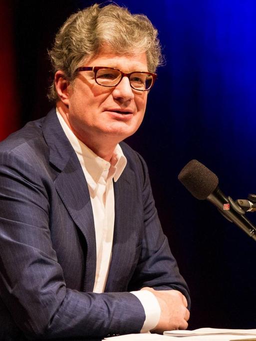 Roger Willemsen sitzt vor einem rotblauem Hintergrund am Mikrofon
