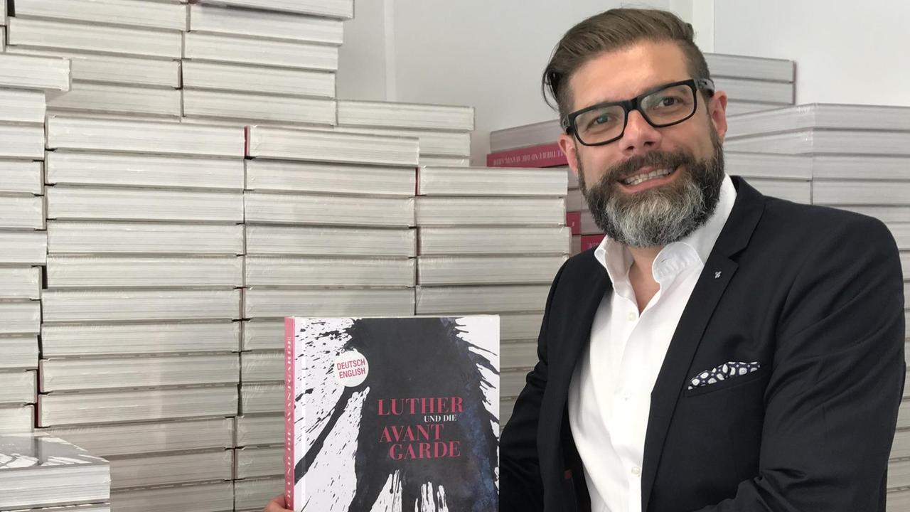 Das Bild zeigt Ulrich Schneider, Geschäftsführer vom "Reformationsjubiläum 2017 e.V.", der vor einem wandhohen Stapel Bücher steht und das Buch mit dem Titel "Luther und die Avantgarde in die Kamera hält.