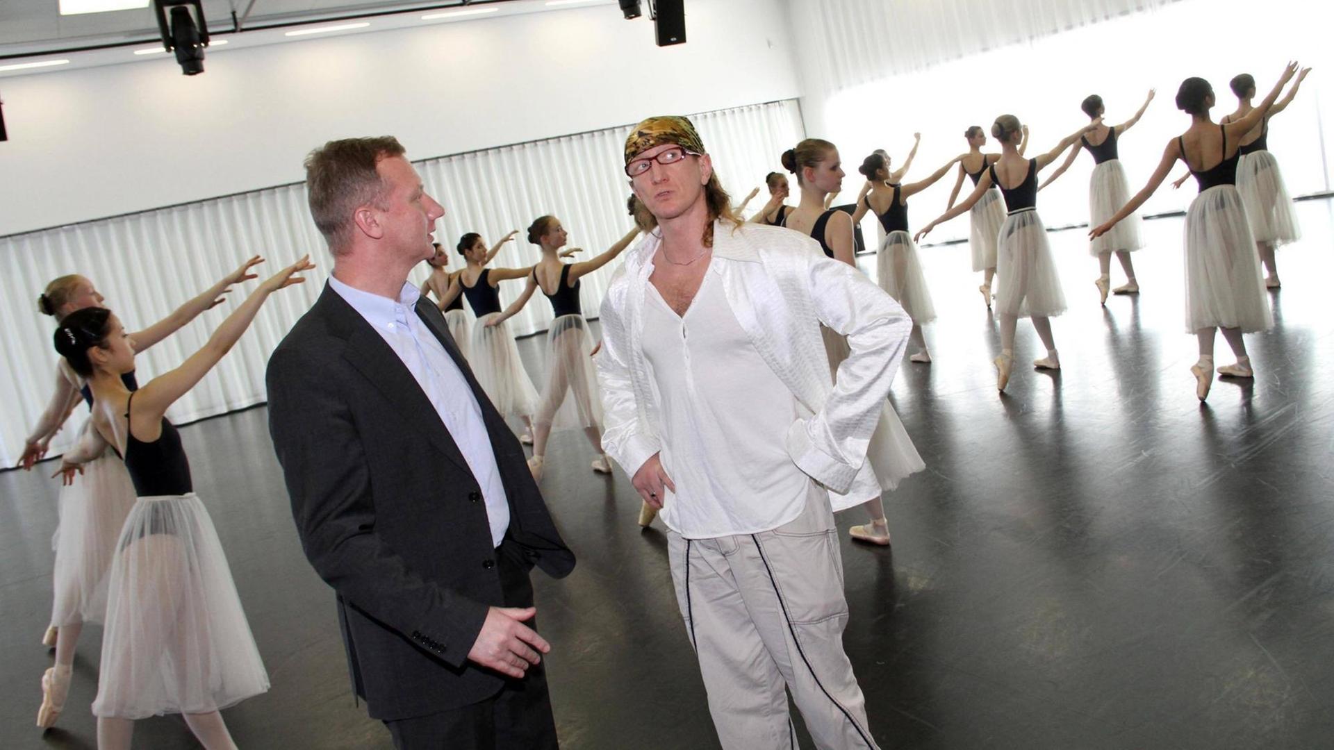 Der ehemalige Schulleiter, Prof. Dr. Ralf Stabel (li.) und der ehemalige künstlerischer Leiter, Prof Gregor Seyffert (re.), in einem Tanzsaal mit einer Gruppe Balletttänzerinnen im Hintergrund an der Staatlichen Ballettschule Berlin.