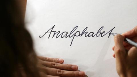 Blick auf ein Blatt Papier, auf den eine Frau das Wort "Analphabet" schreibt.
