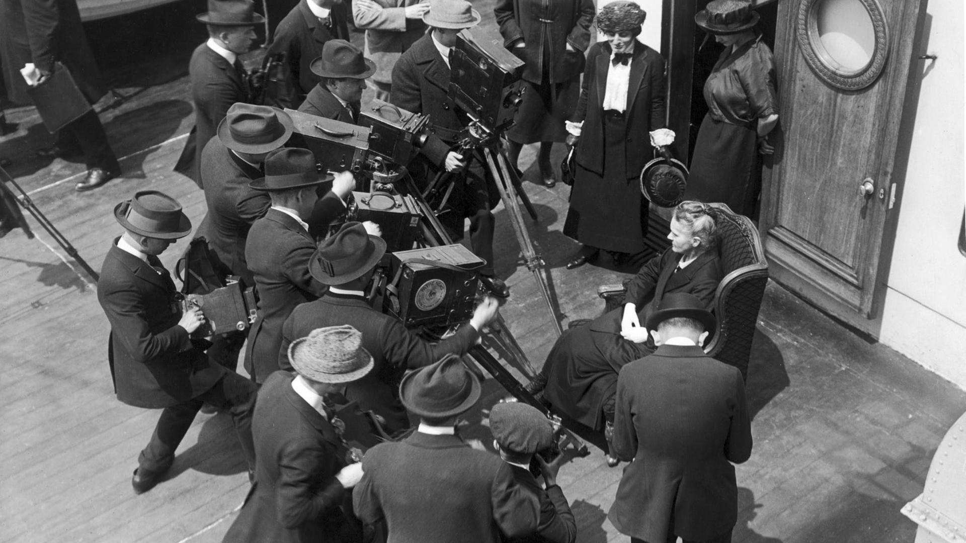Marie Curie auf einem Schiff, umgeben von Kameramännern.