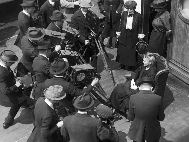 Marie Curie auf einem Schiff, umgeben von Kameramännern.