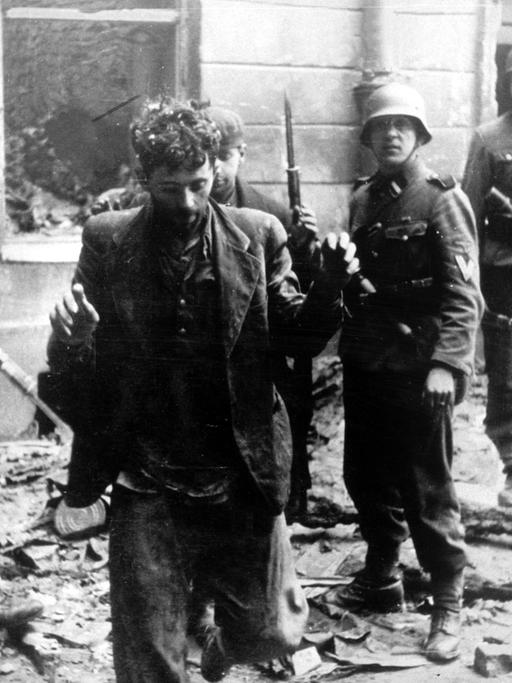 Zwei Juden, die sich in einem Haus versteckt hatten, werden von SS-Soldaten gefangen genommen. Die Aufnahme entstand während des Warschauer Ghetto-Aufstands, der vom 19. April 1943 bis zu seiner blutigen Niederschlagung am 16. Mai 1943 dauerte. Die Nationalsozialisten hatten ein Jahr nach der Besetzung Polens im November 1940 in Warschau ein Ghetto errichtet und dorthin annähernd eine halbe Million Juden verschleppt. Zwischen Juli und September 1942 wurden 300 000 Opfer in den Todeslagern, die meisten in Treblinka, ermordet. Als am 19. April 1943 die SS mit der Verschleppung der restlichen 60 000 Ghetto-Einwohner begann, leisteten mehrere hundert militärisch organisierte Juden bewaffneten Widerstand. Bei den Kämpfen wurden etwa 14 000 jüdische Aufständische getötet.