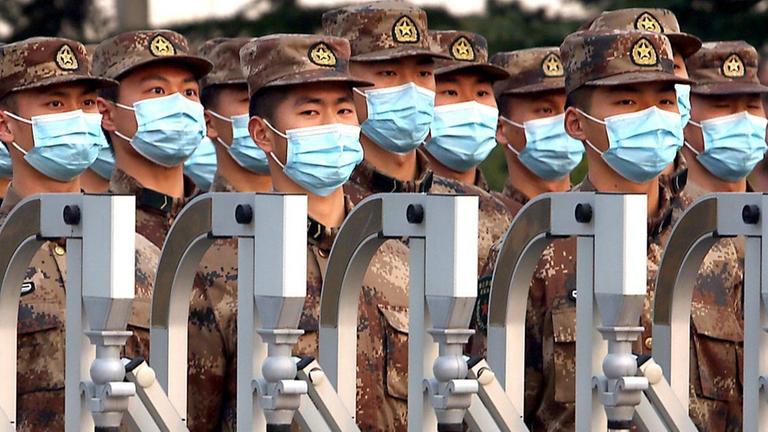 Chinesische Soldaten mit OP-Masken maschieren hinter einem Zaun.