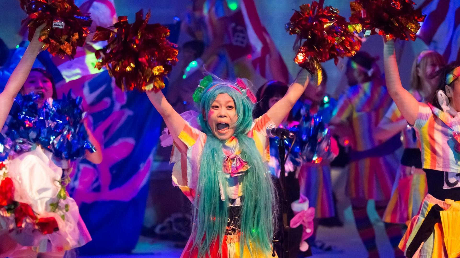 Eine asiatische Frau tanzt mit in die Luft gestreckten Armen in buntem Kostüm und mit bunter Perücke auf einer Bühne.