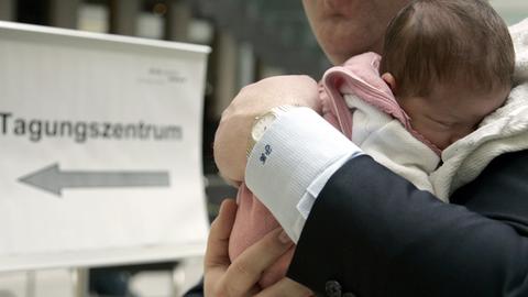 Ein Mann im Anzug trägt ein kleines Baby auf dem Arm, im Hintergrund ist ein Schild mit der Aufschrift "Tagungszentrum" zu sehen.