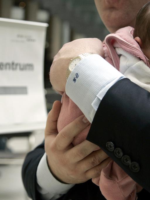 Ein Mann im Anzug trägt ein kleines Baby auf dem Arm, im Hintergrund ist ein Schild mit der Aufschrift "Tagungszentrum" zu sehen.