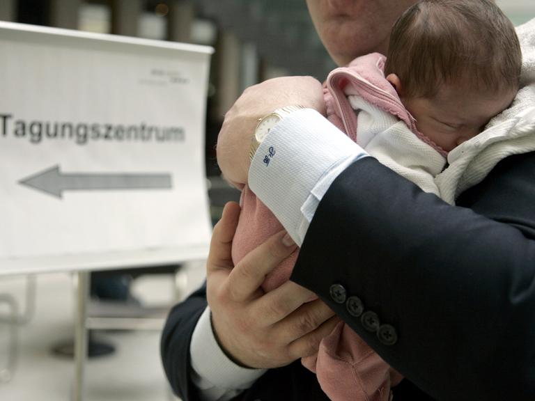 Ein Mann im Anzug trägt ein kleines Baby auf dem Arm, im Hintergrund ist ein Schild mit der Aufschrift "Tagungszentrum" zu sehen.