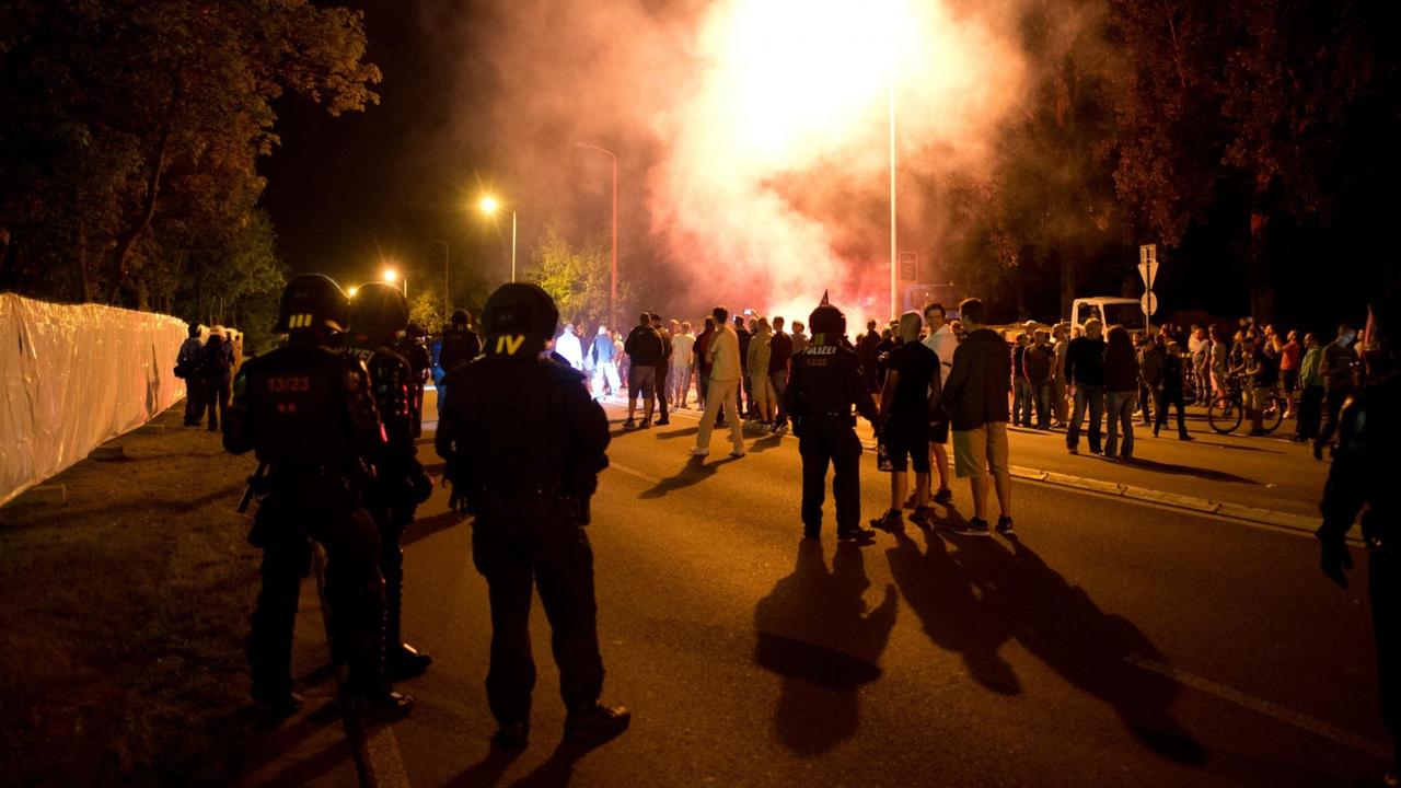 Demonstranten und Polizisten in Schutzkleidung.stehen in der Nacht auf einer Straße. Im Hintergrund sind Leuchtfeuer und Rauch zu sehen.