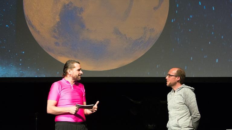 Astronom Dr. Florian Freistetter auf der Bühne im Gespräch mit Martin Putingham. Im Hintergrund ist ein Mond auf die Leinwand projiziert.