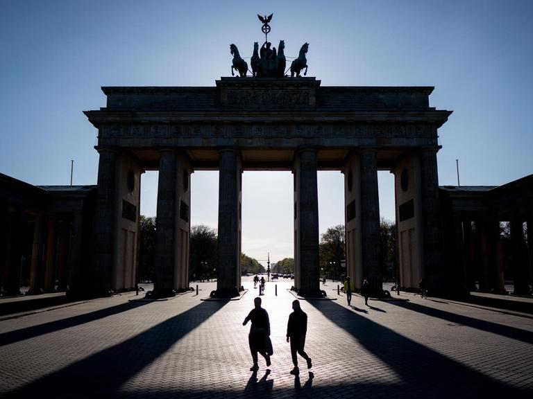 Das Brandenburger Tor in Berlin wirft in der untergehenden Sonne lange Schatten, zwei Personen gehen mittig davor entlang.