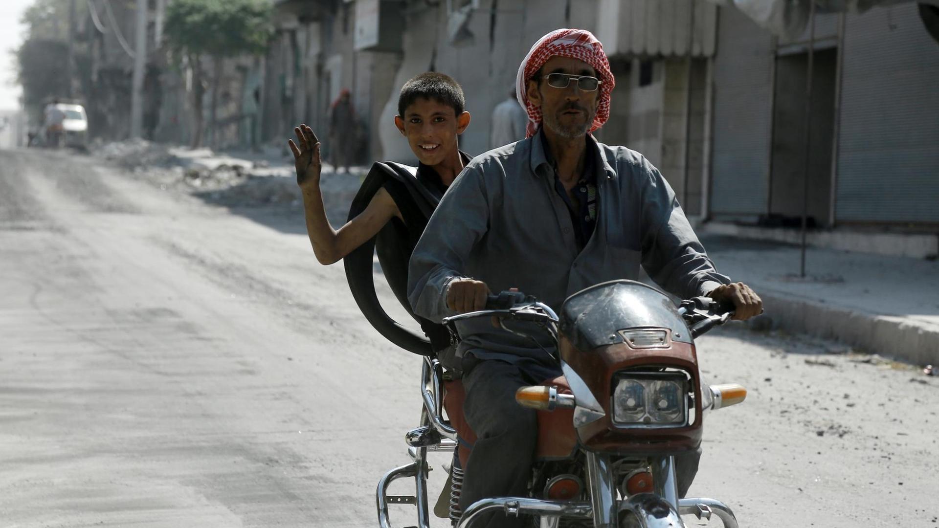 Ein Mann und ein Junge fahren auf einem Motorroller durch eine Straße in Manbidsch, der Junge winkt in die Kamera.