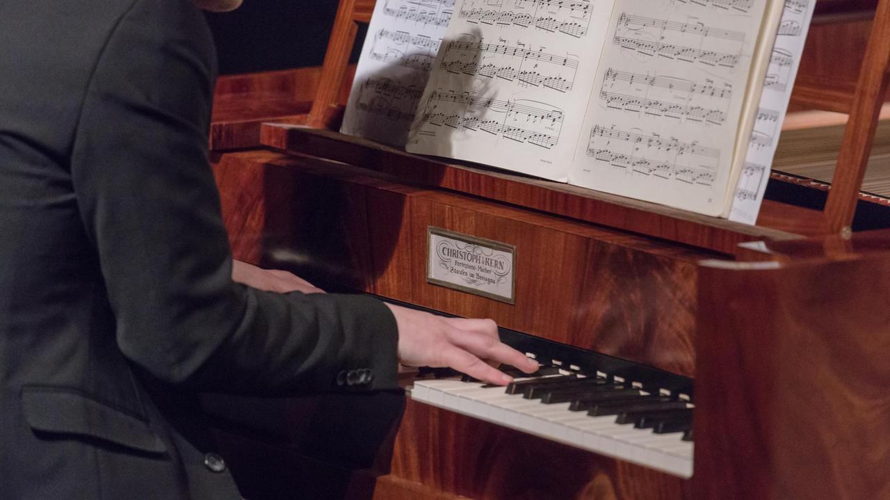 An einem holzfarbenen und -gemaserten hammerflügel sieht man die Hände eines Mannes in schwarzem Anzug auf der Klaviatur liegen. Ein Schild am Instrument zeigt den Namen des Klavierbauers, auf dem Notenpult stehen aufgeschlagene Noten. 