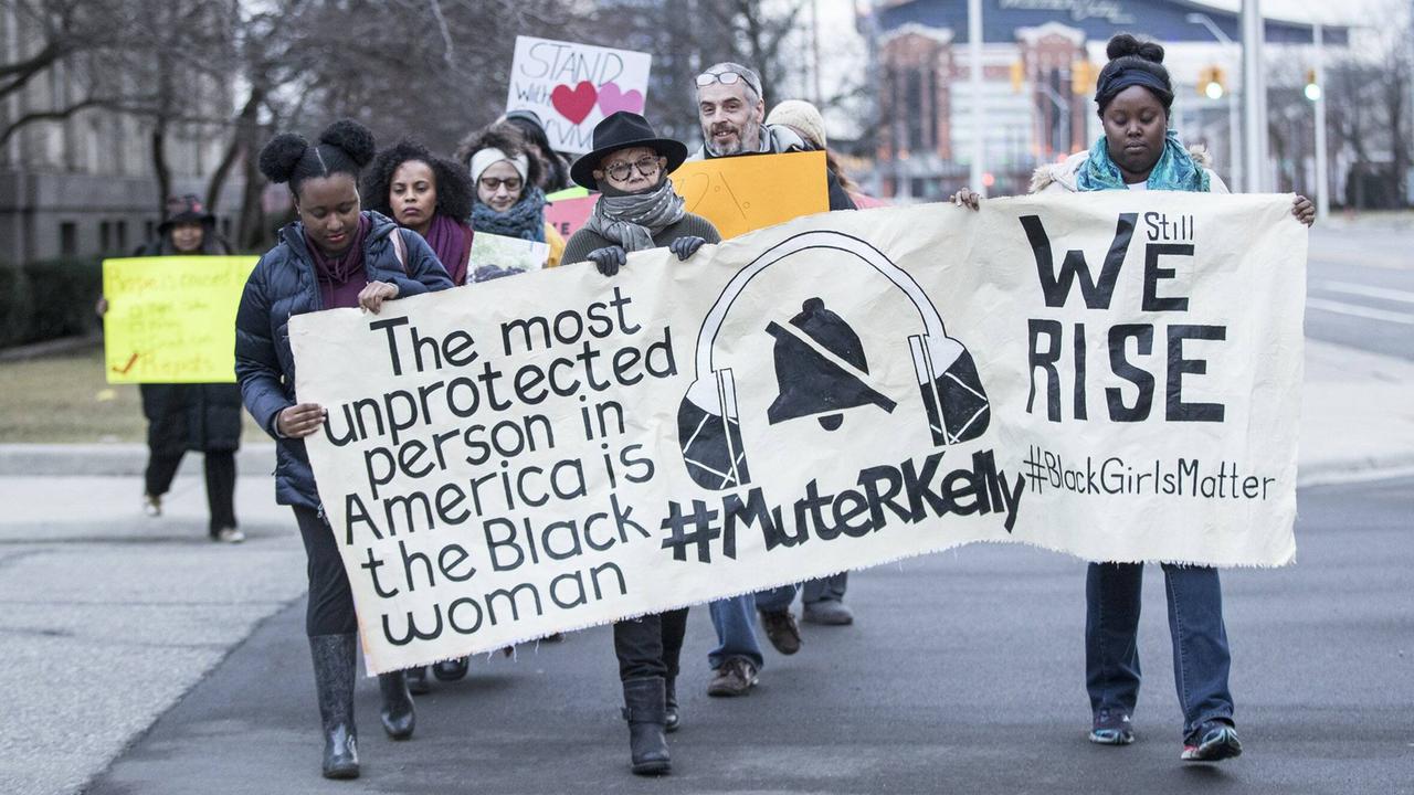 Mehrere Frauen gehen mit einem Plakat in den Händen, auf dem "#MuteRKelly" in der Mitte steht.