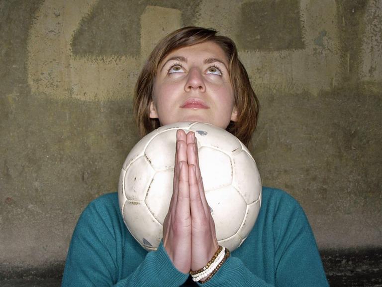 Eine junge Frau mit Fußball und gefalteten Händen hat ihren Blick nach oben gerichtet.