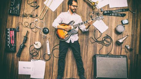 Das Bild zeigt einen rücklings auf dem Boden liegenden, Gitarre spielenden jungen Mann aus der Vogelperspektive. Um ihn herum liegt Musik-Equipement.