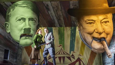 Die Opernsänger Barbara Senator und Santiago Sanchez in Kostümen auf der Bühne am Theater Bonn. Hinter ihnen sind Porträts von Adolf Hitler und Winston Churchill als Figuren mit Klappmaul zu sehen.