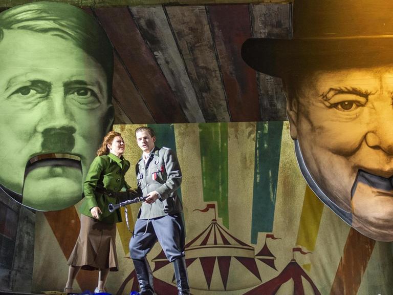 Die Opernsänger Barbara Senator und Santiago Sanchez in Kostümen auf der Bühne am Theater Bonn. Hinter ihnen sind Porträts von Adolf Hitler und Winston Churchill als Figuren mit Klappmaul zu sehen.