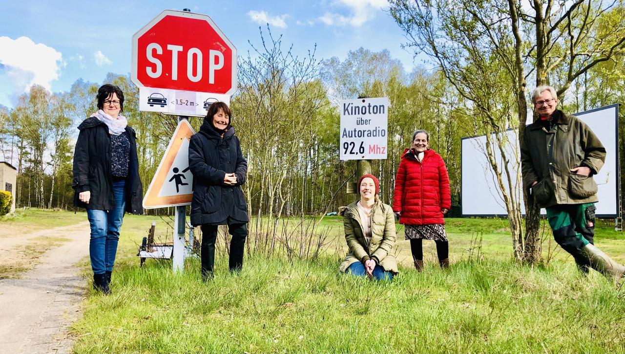 Fünf Menschen vor einem Schild im einer Wiesen- und Waldlandschaft. Auf einem Schild steht "Kinoton über Autoradio 92,6 Mhz"