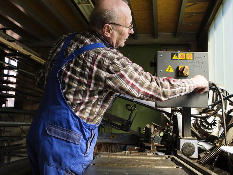 Ein älterer Mann in kariertem Hemd und blauer Arbeitshose bedient eine Maschine in einem mittelständischem Unternehmen
