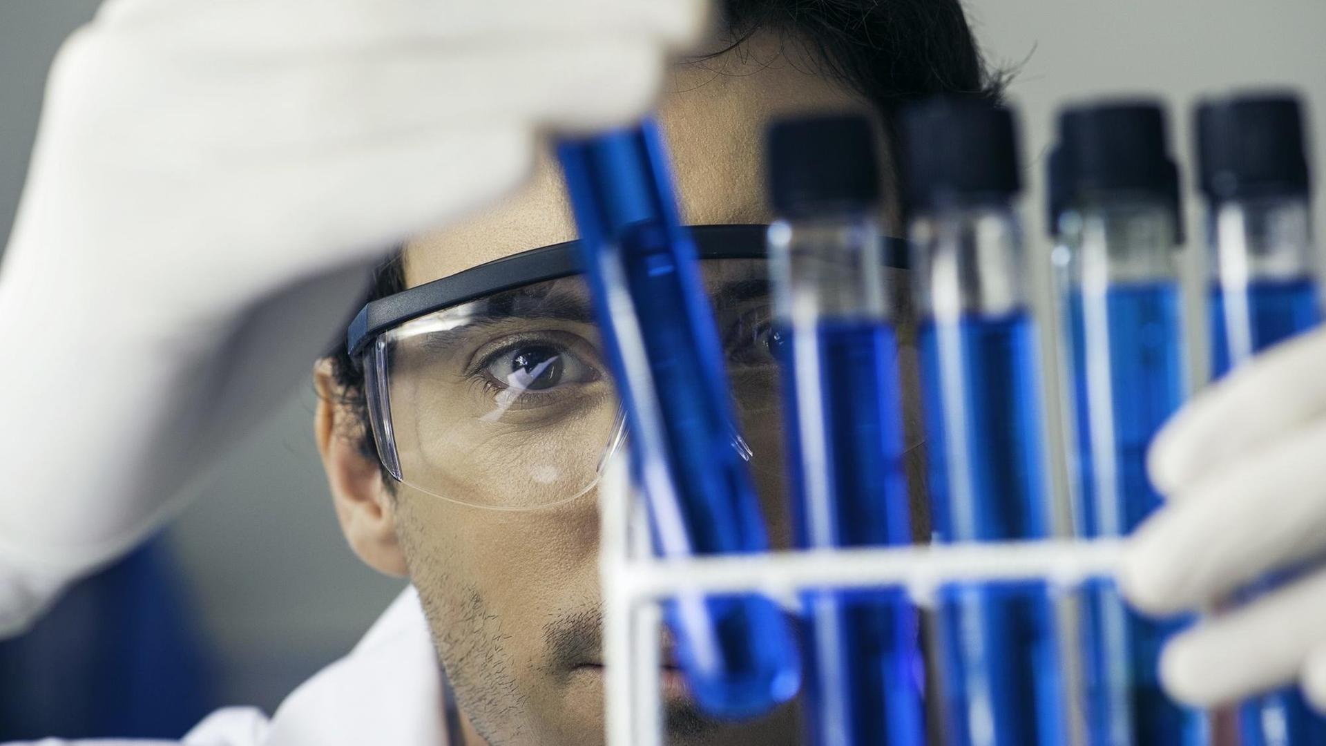 Ein Mann mit weißem Kittel und Schutzbrille vor einem Ständer mit Reagenzgläsern in einem Labor.