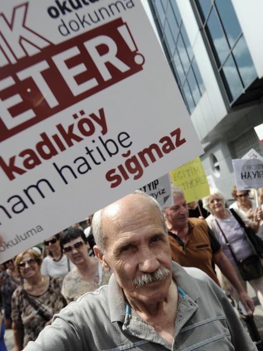 Ein Mann hält ein Protestplakat in die Kamera auf dem auf Türkisch steht: "Genug! Hört auf, unsere Schulen in Imam Hatips zu verwandeln!", im Hintergrund gehen auf einer Straße weitere Demonstranten.