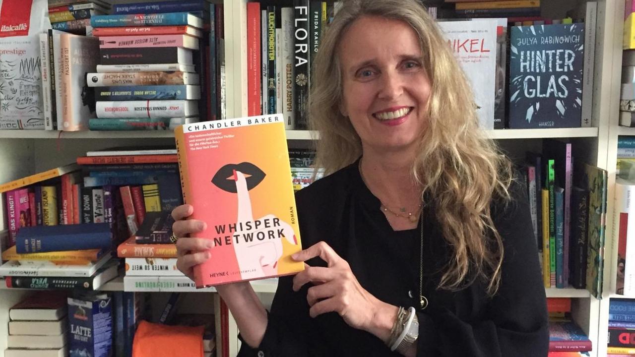 Lesart-Redakteurin Kim Kindermann steht mit dem Buch "Whisper Network" von Chandler Baker vor einem Bücherregal.
