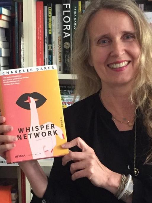 Lesart-Redakteurin Kim Kindermann steht mit dem Buch "Whisper Network" von Chandler Baker vor einem Bücherregal.