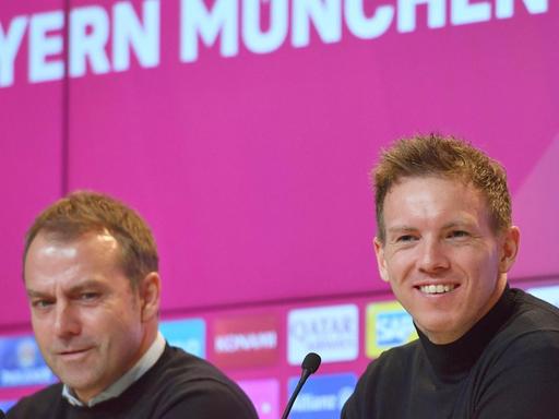 Zwei Männer sitzen in einer Pressekonferenz. Hinter ihnen ist ein magenta farbener Hintergrund auf dem Bayern München steht. Es sind Hansi Flick und sein Nachfolger Julian Nagelsmann.