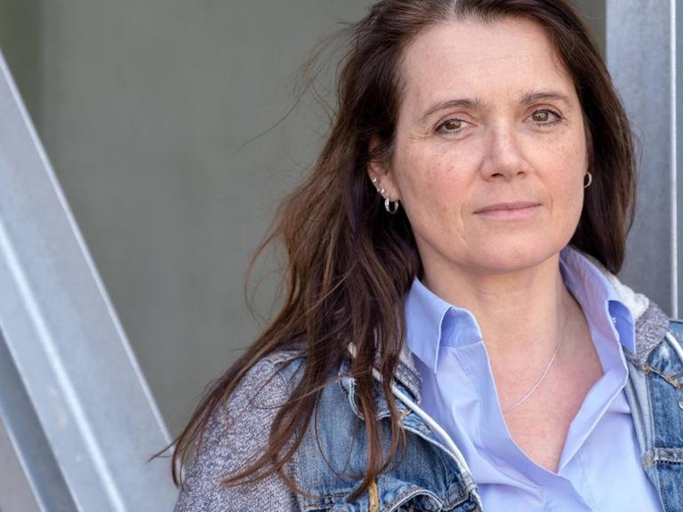 Barbara Junge steht in legerer Kleidung und mit ernstem Blick vor einer Metallkonstruktion.