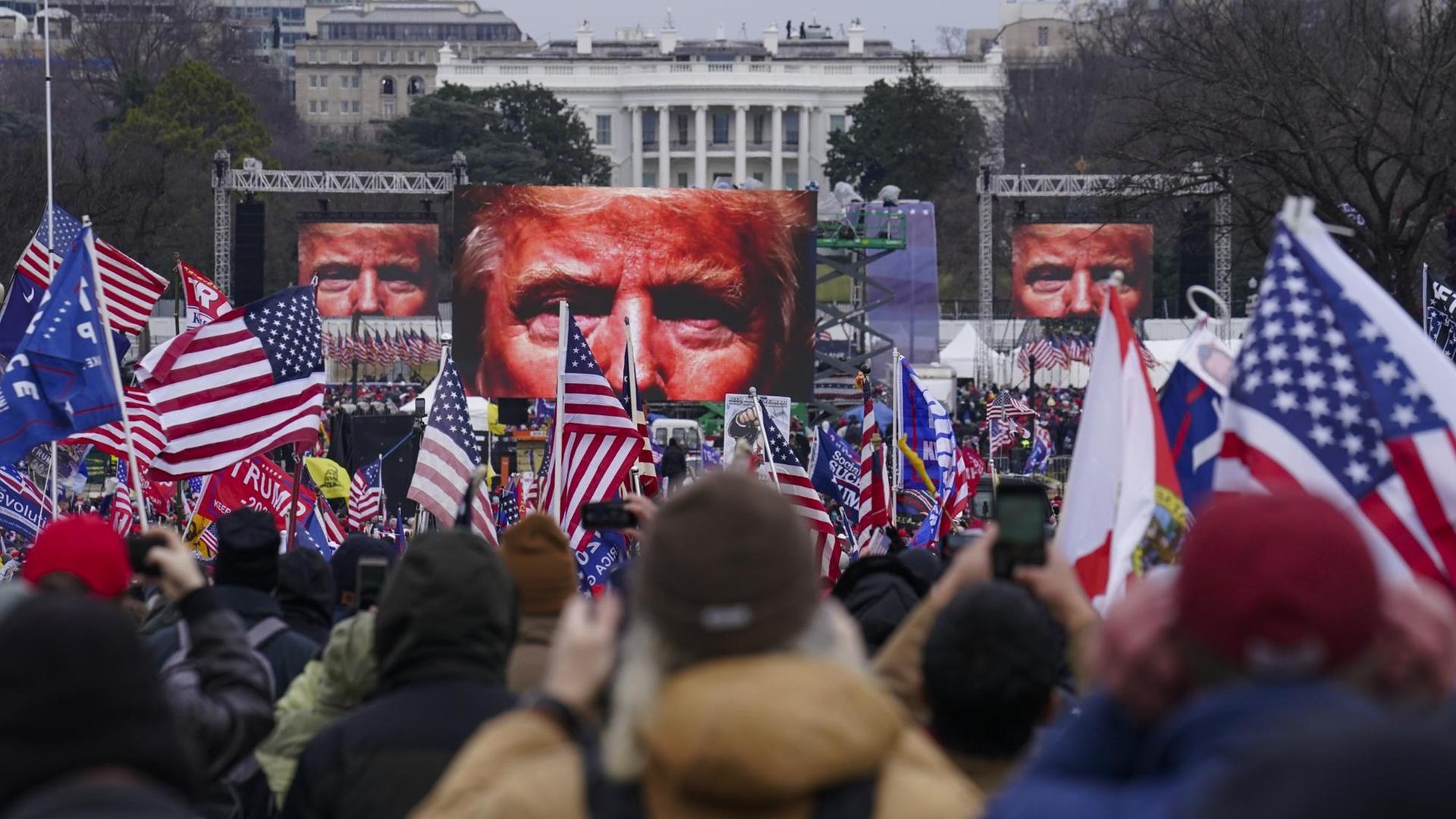 Auf dem Bild sind Demonstranten vor dem weißen Haus zu sehen. Vor ihnen stehen Bildschirme, die das Gesicht von Donald Trump zeigen.