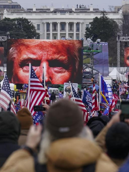 Auf dem Bild sind Demonstranten vor dem weißen Haus zu sehen. Vor ihnen stehen Bildschirme, die das Gesicht von Donald Trump zeigen.
