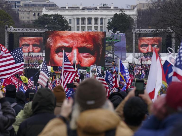 Auf dem Bild sind Demonstranten vor dem weißen Haus zu sehen. Vor ihnen stehen Bildschirme, die das Gesicht von Donald Trump zeigen.