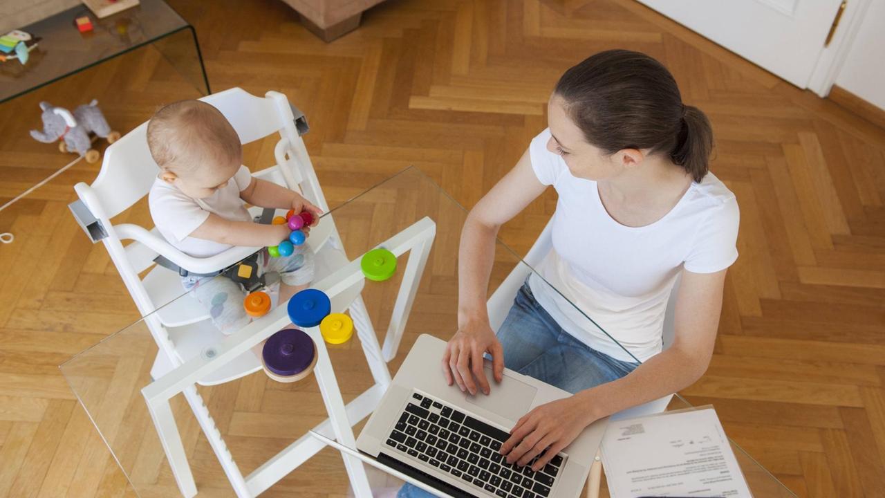 Eine Frau sitzt neben einem ein Baby im Hochstuhl am Tisch vor einem Laptop.