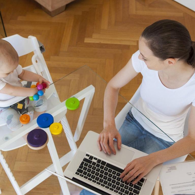 Eine Frau sitzt neben einem ein Baby im Hochstul am Tisch vor einem Laptop