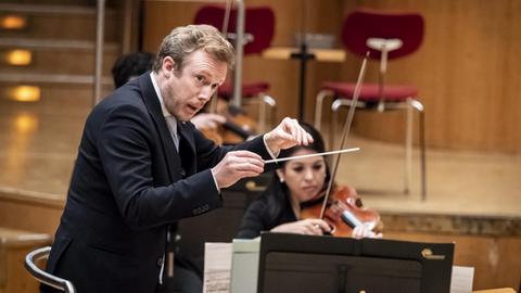 Ein dirigierender Mann steht auf einem Podest und blickt zur Seite. Vom Orchester ist nur eine Geigerin zu erkennen.